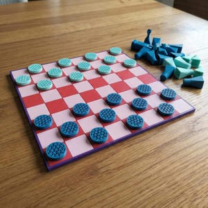 K&ouml;nnte beinhalten: Ein Dame-Spielset mit einem rosa-rot karierten Brett und blauen und t&uuml;rkisfarbenen Spielsteinen. Zus&auml;tzliche blaue Spielsteine sind in der N&auml;he verstreut. Das Brett ist lila umrandet und liegt auf einer Holzoberfl&auml;che.