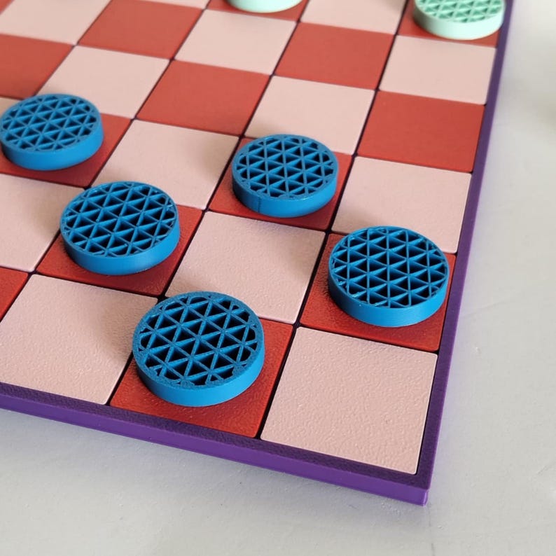 K&ouml;nnte beinhalten: Nahaufnahme eines Schachbretts mit blauen und gr&uuml;nen Schachfiguren. Das Brett hat ein rot-rosa kariertes Muster mit einem lila Rand. Die blauen Schachfiguren haben ein geometrisches Design.