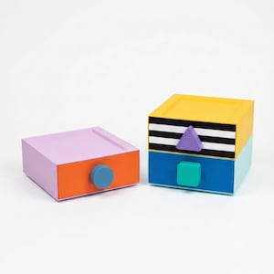 Könnte beinhalten: Zwei farbenfrohe Dekorationsboxen. Eine ist lavendelfarben mit einer orangefarbenen Vorderseite und einem blauen runden Knopf. Die andere ist gelb, schwarz, weiß, blau und blaugrün mit einem lila Dreieck und einem türkisfarbenen quadratischen Knopf. Beide haben eine Schublade.