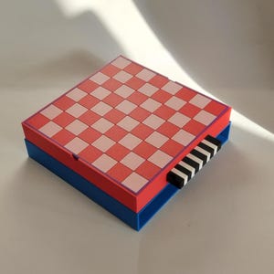 K&ouml;nnte beinhalten: Ein farbenfrohes, geometrisches Schachspiel. Das Schachbrett ist rot und wei&szlig; kariert, mit einem roten Rand, und sitzt auf einer blauen Basis. Die Schachfiguren sind schwarz und wei&szlig;. Das Set ist kompakt und modern.