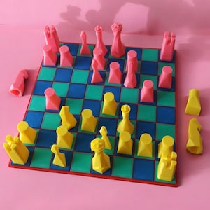 Op de afbeelding: Een geometrisch schaakspel met roze en gele stukken op een blauw, groen en rood geblokt bord. De stukken zijn klaar om te spelen. De achtergrond is effen roze.