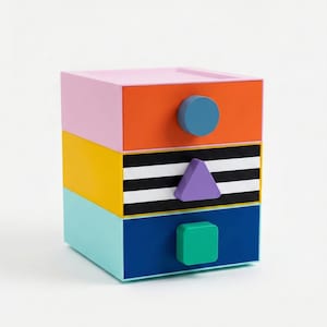 May include: A multi-coloured three-drawer storage unit. The top drawer is pink, the middle is orange, and the bottom is yellow. Each drawer has a different shaped knob: a circle, a triangle, and a square. The unit is approximately 25 cm wide, 25 cm deep, and 25 cm tall.
