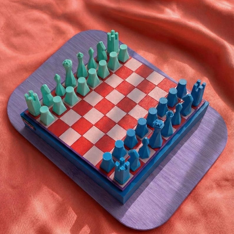 K&ouml;nnte beinhalten: Ein farbenfrohes Schachspiel mit einem rot-wei&szlig; karierten Brett. Die Schachfiguren sind t&uuml;rkis und blau, angeordnet auf einer lila Basis. Das Set wird auf einem roten Stoffhintergrund pr&auml;sentiert.