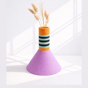 May include: A contemporary vase featuring a lavender cone base and an orange cylindrical neck. The neck has a band of black and teal stripes. The vase holds dried ornamental grass. A decorative home accent, the vase is a striking design.