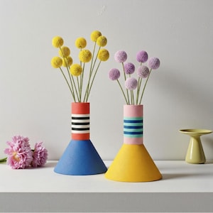 May include: Two vibrant vases with flowers. One is blue and red with black and white stripes, holding yellow flowers. The other is yellow and pink with blue stripes, holding purple flowers. A small yellow side table is in the background.