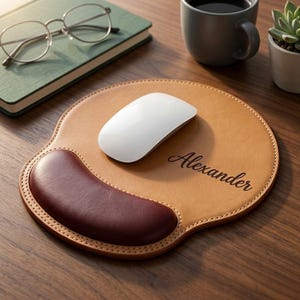 May include: A tan leather mouse pad with a burgundy wrist rest and the name "Alexander" in script. A white wireless mouse sits on the pad. The pad is on a wooden desk, next to a cup of coffee and a succulent.