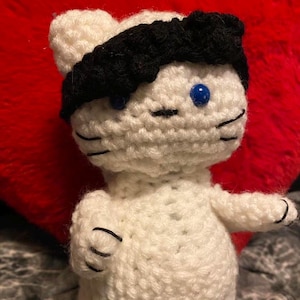 Puede incluir: Muñeca de gato de ganchillo blanco hecha a mano con una diadema negra y ojos azules de botón. El gato tiene detalles negros cosidos para la nariz, la boca y los bigotes. La muñeca está sobre un fondo rojo en forma de corazón.