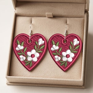 May include: A pair of heart-shaped drop earrings featuring a deep red embroidered base with a textured pattern. Each earring displays a white floral design with green leaves and stems, finished with silver-toned metal hooks for attachment.