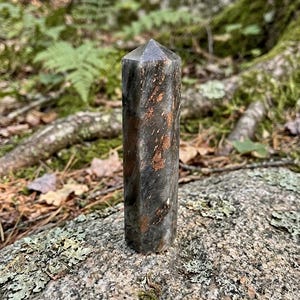 Könnte beinhalten: Ein polierter, sechseckiger Kristallturm mit einer dunkelgrauen und schwarzen Basis, der durchgehend metallische, kupferfarbene Einschlüsse auf der Steinoberfläche aufweist und in einer pyramidenförmigen Spitze endet.