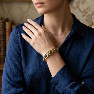 Puede incluir: Una pulsera de eslabones en tono dorado que presenta una alternancia de esferas metálicas pulidas y segmentos rectangulares texturizados. La pulsera se lleva en la muñeca, mostrando un patrón geométrico repetitivo con un acabado cálido y brillante.
