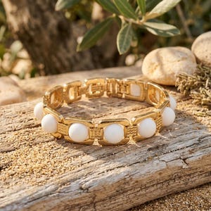 Puede incluir: Una pulsera de eslabones en tono dorado que presenta una serie de cuentas blancas redondas y lisas engastadas en marcos metálicos rectangulares. La pulsera está construida con segmentos articulados y un cierre seguro, con acabado metálico pulido.