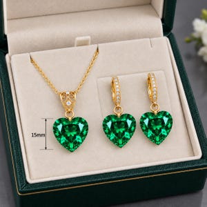 Può includere: Un set di gioielli color oro composto da una collana e due orecchini a cerchio, ciascuno decorato con una pietra preziosa verde a forma di cuore. Il ciondolo misura 15 mm di altezza. Gli orecchini sono impreziositi da piccole pietre chiare.