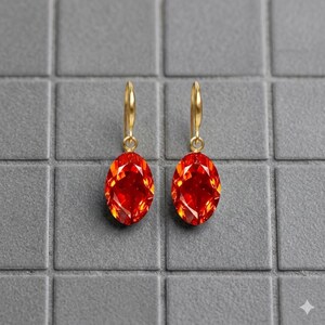 Puede incluir: Un par de pendientes colgantes dorados con grandes piedras preciosas rojas ovaladas y facetadas. Los pendientes cuelgan de un diseño de gancho simple. Las piedras preciosas tienen una apariencia brillante y ardiente sobre un fondo gris.