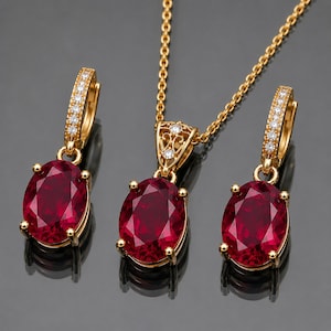 Könnte beinhalten: Ein Goldschmuckset bestehend aus einer Anhängerkette und zwei passenden Creolen. Jedes Schmuckstück verfügt über einen großen, oval geschliffenen, tiefroten Edelstein, der von vier Krappen gehalten wird, sowie kleine klare Steine am Gold.