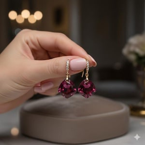 Puede incluir: Un par de pendientes dorados con piedras preciosas cuadradas de color rojo intenso. Los pendientes tienen un pequeño aro con diamantes. Las piedras preciosas están facetadas. Una mano sostiene los pendientes.