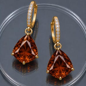 May include: A pair of gold-colored hoop earrings with a row of small, clear stones along the top. Each earring has a large, triangular, orange-brown gemstone pendant with gold-colored accents at the corners. The earrings are displayed on a clear surface.