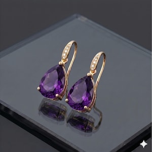 Può includere: Un paio di orecchini pendenti color oro con grandi pietre preziose viola a forma di pera. La parte superiore di ogni orecchino presenta un design curvo con piccole pietre trasparenti. Gli orecchini sono esposti su una superficie riflettente.