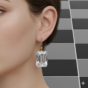 Op de afbeelding: Een gouden oorbel met een grote, heldere, rechthoekige edelsteen. De oorbel hangt aan het oor van een persoon. De edelsteen is facetgeslepen en reflecteert licht. De achtergrond is een patroon van zwarte, witte en grijze tegels.