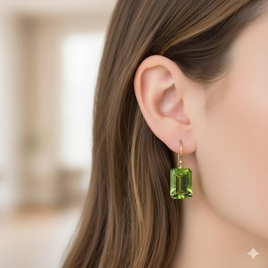 Può includere: Primo piano di un orecchino dorato con una gemma rettangolare verde chiaro. L'orecchino è appeso all'orecchio di una persona, con l'attenzione focalizzata sul gioiello. La gemma è di un colore vivace e accattivante.