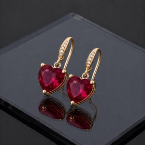 Puede incluir: Un par de pendientes colgantes de oro con piedras preciosas rojas en forma de corazón. Cada pendiente tiene un gancho curvo engastado con pequeñas piedras claras que conduce a un colgante rojo vibrante y facetado en forma de corazón.