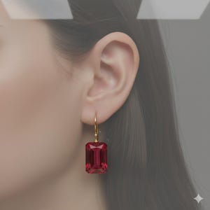 K&ouml;nnte beinhalten: Goldfarbener Ohrring mit einem rechteckigen, tiefroten Edelstein. Der Edelstein ist facettiert und f&auml;ngt das Licht ein. Der Ohrring ist im Tropfenstil gehalten und h&auml;ngt an einem einfachen Haken.