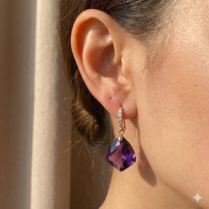 May include: A close-up of a rose gold earring with a large, faceted, deep purple amethyst gemstone. The earring has a small, sparkling diamond-studded hoop. The earring is dangling from a person's ear.