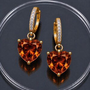 May include: A pair of gold hoop earrings featuring a row of small clear stones along the front and a large, faceted, heart-shaped orange gemstone dangling from each hoop. The earrings are crafted with a polished gold finish and secure clasps.