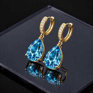 May include: A pair of gold hoop earrings featuring a row of small clear stones along the front of the hoop and a large, faceted, pear-shaped blue gemstone dangling from each earring. The metal has a polished gold finish and a hinged closure.