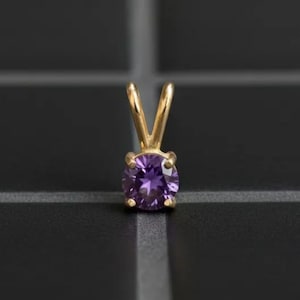 May include: A gold pendant with a round, deep purple gemstone. The gemstone is held in place by a gold setting with a V-shaped bail. The pendant is set against a dark, textured background.