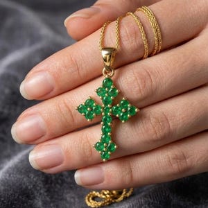 Puede incluir: Un collar con colgante de cruz dorada y cadena fina. La cruz está adornada con numerosas piedras preciosas verdes. El collar se muestra sobre un fondo gris suave, resaltando los detalles de la joya.