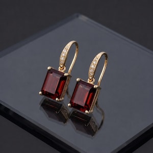 Peut inclure: Une paire de boucles d'oreilles dorées avec des pierres précieuses rectangulaires rouge foncé. Les boucles d'oreilles ont un design à crochet et sont ornées de petites pierres claires sur la courbe supérieure. Les boucles d'oreilles sont présentées sur une surface réfléchissante.