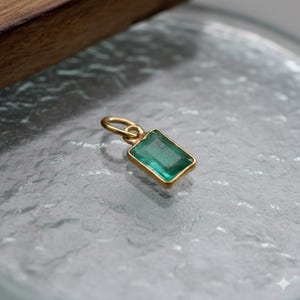 Könnte beinhalten: Ein rechteckiger Anhänger im Smaragdschliff mit einem leuchtend grünen Edelstein, der in einer goldfarbenen Lünette gefasst ist. Der Anhänger hat eine kleine Schlaufe zum Anbringen an einer Kette. Der Schmuck wird auf einer strukturierten, hellen Oberfläche präsentiert.