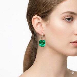 Op de afbeelding: Een goudkleurige oorbel met een grote, vierkant geslepen, smaragdgroene edelsteen. De oorbel heeft een klein, met diamanten bezet accent boven de groene steen. Op een witte achtergrond.