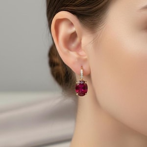 Peut inclure: Boucles d'oreilles pendantes dorées avec une grosse pierre précieuse rouge foncé de coupe ovale. La partie supérieure de la boucle d'oreille présente un petit motif incurvé avec des pierres d'accent claires. Les boucles d'oreilles sont portées.