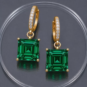 May include: A pair of gold hoop earrings featuring a row of small clear stones along the front of the hoop and a large, square-cut, deep green gemstone pendant suspended from each hoop by a small gold ring, creating a dangling earring design.
