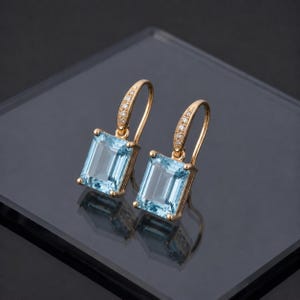 Può includere: Un paio di orecchini dorati con pietre preziose rettangolari azzurro chiaro. Gli orecchini hanno un design a gancio con piccole pietre trasparenti incastonate lungo la curva. Le pietre sono incastonate in una montatura dorata.