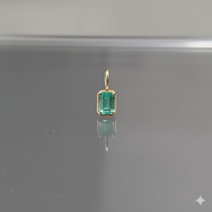 Puede incluir: Un colgante rectangular con esmeralda y una vibrante piedra preciosa verde. El colgante está engastado en un marco dorado con un borde texturizado y un lazo dorado a juego para una cadena. El diseño es simple y elegante.