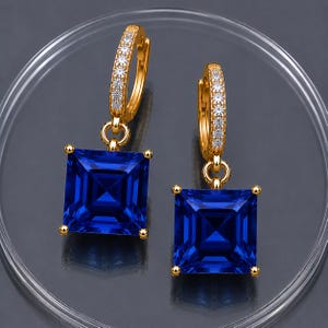 Könnte beinhalten: Ein Paar goldene Creolen mit einer Reihe kleiner, klarer Steine auf der Vorderseite und einem großen, quadratisch geschliffenen, tiefblauen Edelstein, der an jeder Creole hängt. Die quadratischen blauen Steine werden von vier goldenen Krappen gehalten.