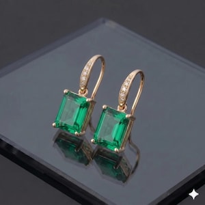 Könnte beinhalten: Ein Paar goldfarbene Ohrringe mit smaragdgrünen Edelsteinen im Smaragdschliff. Die Ohrringe haben einen Klappverschluss und sind mit kleinen, klaren Kristallen verziert.
