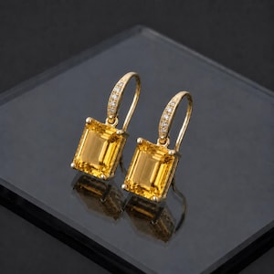 Peut inclure: Une paire de boucles d'oreilles en or avec des pierres précieuses rectangulaires jaunes. Les boucles d'oreilles ont un design en forme de crochet incurvé avec de petites pierres claires serties sur le dessus. Les pierres précieuses sont d'un jaune vif.