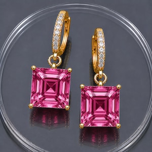 May include: A pair of gold hoop earrings featuring a row of small clear stones along the front and a large, square-cut, vibrant pink gemstone dangling from each hoop. The earrings are designed with a hinged closure mechanism for secure wear.