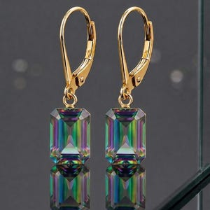 May include: A pair of gold-colored leverback earrings. Each earring features two rectangular, iridescent, multi-colored stones. The stones reflect hues of green, blue, purple, and yellow. The earrings are displayed against a dark background.