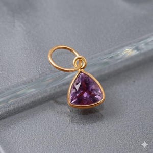May include: A gold pendant featuring a triangular, deep purple gemstone. The gemstone is set in a gold bezel and attached to a small gold loop. The pendant is shown against a grey background.