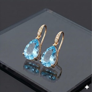 Puede incluir: Un par de pendientes colgantes dorados con piedras preciosas azul claro en forma de pera. Los pendientes tienen un diseño de gancho con pequeñas piedras transparentes engastadas a lo largo de la curva superior.