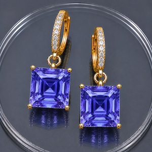 May include: A pair of gold hoop earrings featuring a row of small clear stones along the front and a large, square-cut, vibrant purple gemstone dangling from each hoop. The earrings have a polished gold finish and a secure hinged closure mechanism.