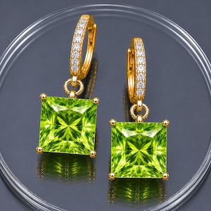 May include: A pair of gold hoop earrings featuring a square-cut, vibrant lime green gemstone pendant. The hoop portion of each earring is embellished with a row of small, clear, sparkling stones, and the square gems are held by four prongs.