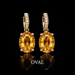 May include: A pair of gold hoop earrings featuring oval-cut yellow gemstones suspended from the hoops. The hoops are accented with small, clear, round-cut stones along the front. The word OVAL is printed in white text below the earrings.