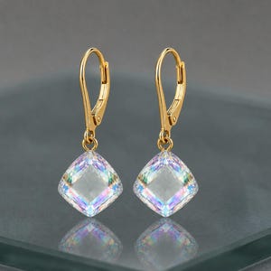 May include: A pair of gold-colored leverback earrings. Each earring features a square, iridescent crystal pendant. The crystals reflect a spectrum of colors, including pink, blue, and green, creating a shimmering effect. The earrings are displayed on a reflective surface.