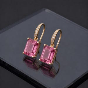 May include: A pair of gold drop earrings featuring rectangular, pink gemstones. The earrings have a curved top adorned with small, clear stones. The earrings are presented on a reflective surface.
