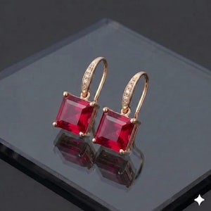 Peut inclure: Une paire de boucles d'oreilles dorées avec des pierres précieuses carrées rouge foncé. Les boucles d'oreilles ont un design en forme de crochet avec de petites pierres d'accent claires. Les boucles d'oreilles sont présentées sur une surface réfléchissante.